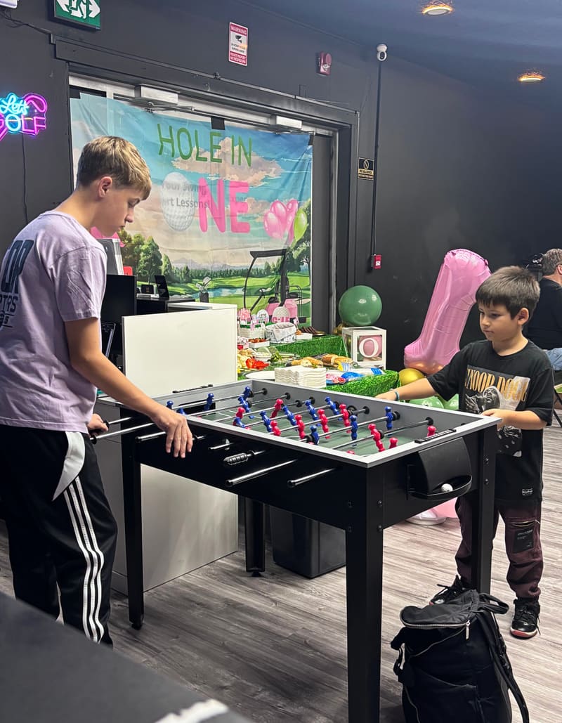 Kids' birthday party at Club X Indoor Golf — children at the foosball table with a HOLE IN ONE banner backdrop